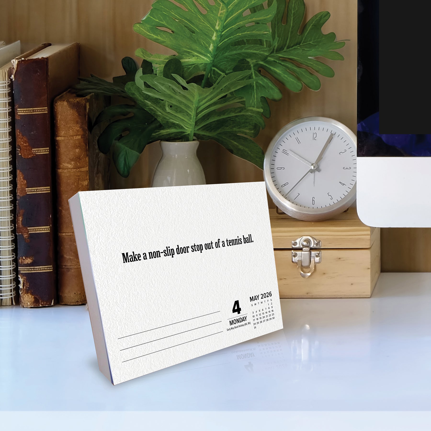 2026 Daily Box Calendar with life hack tip on a desk next to books, a clock, and a plant.