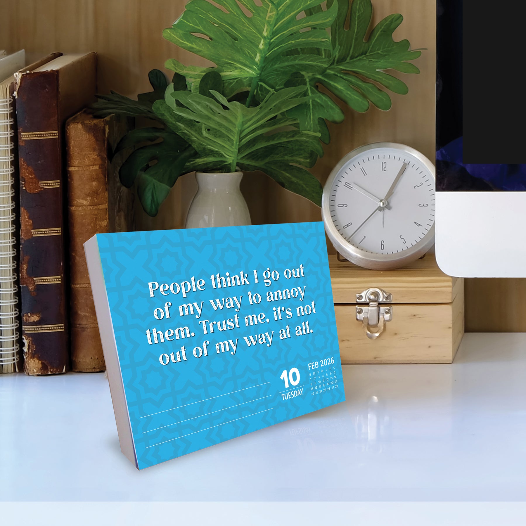 2026 Daily Box Calendar featuring humorous quote and vibrant blue design on a desk with plants and clock.