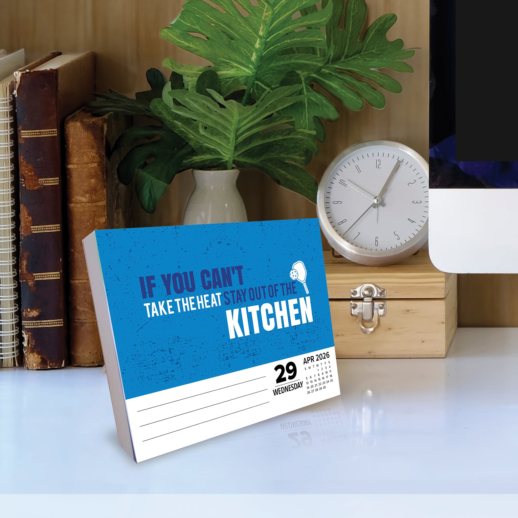 2026 Daily Box Calendar featuring kitchen humor and vibrant design on a desk with plants and books.