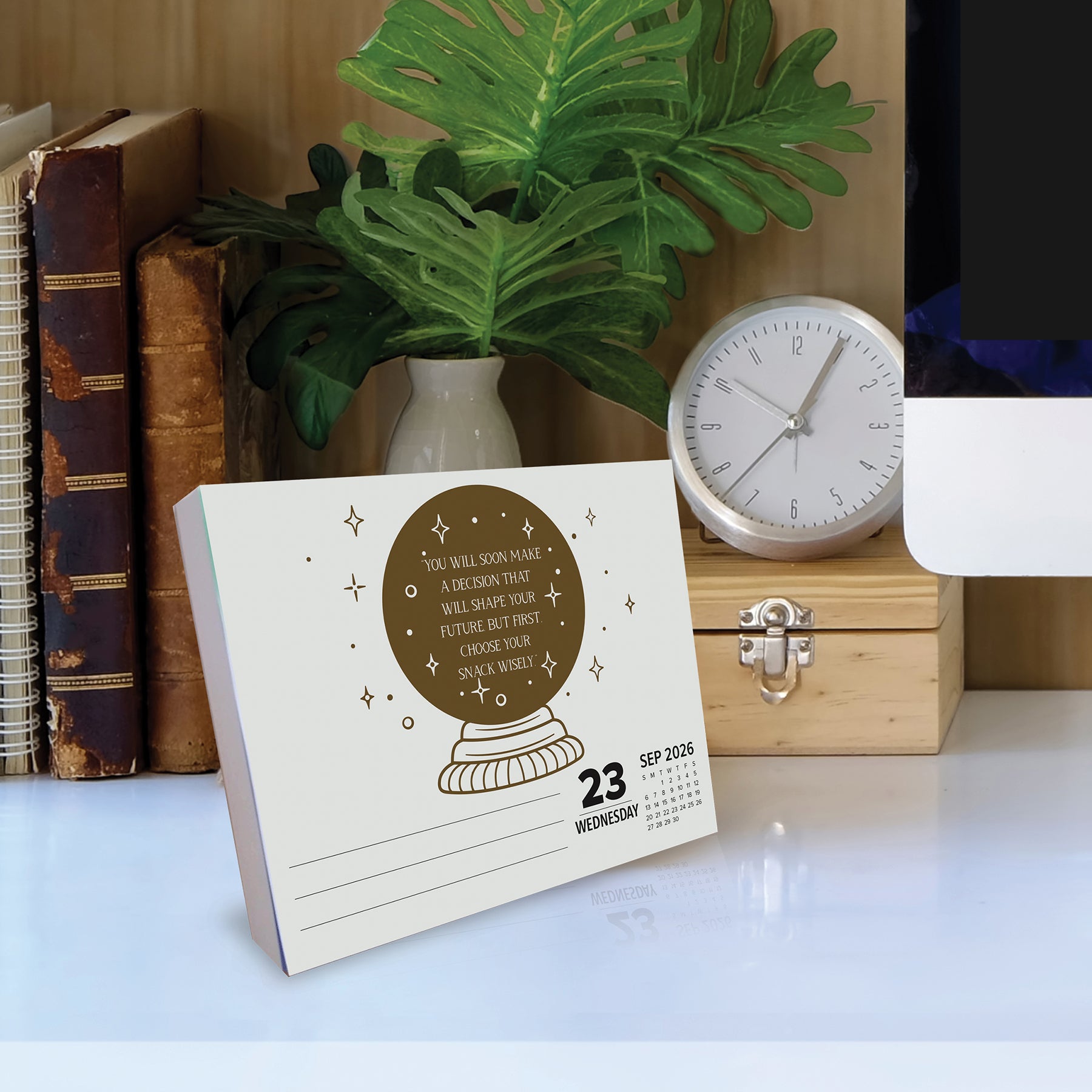 Miss Fortune 2026 Daily Box Calendar with humorous predictions on a desk, surrounded by plants and books.